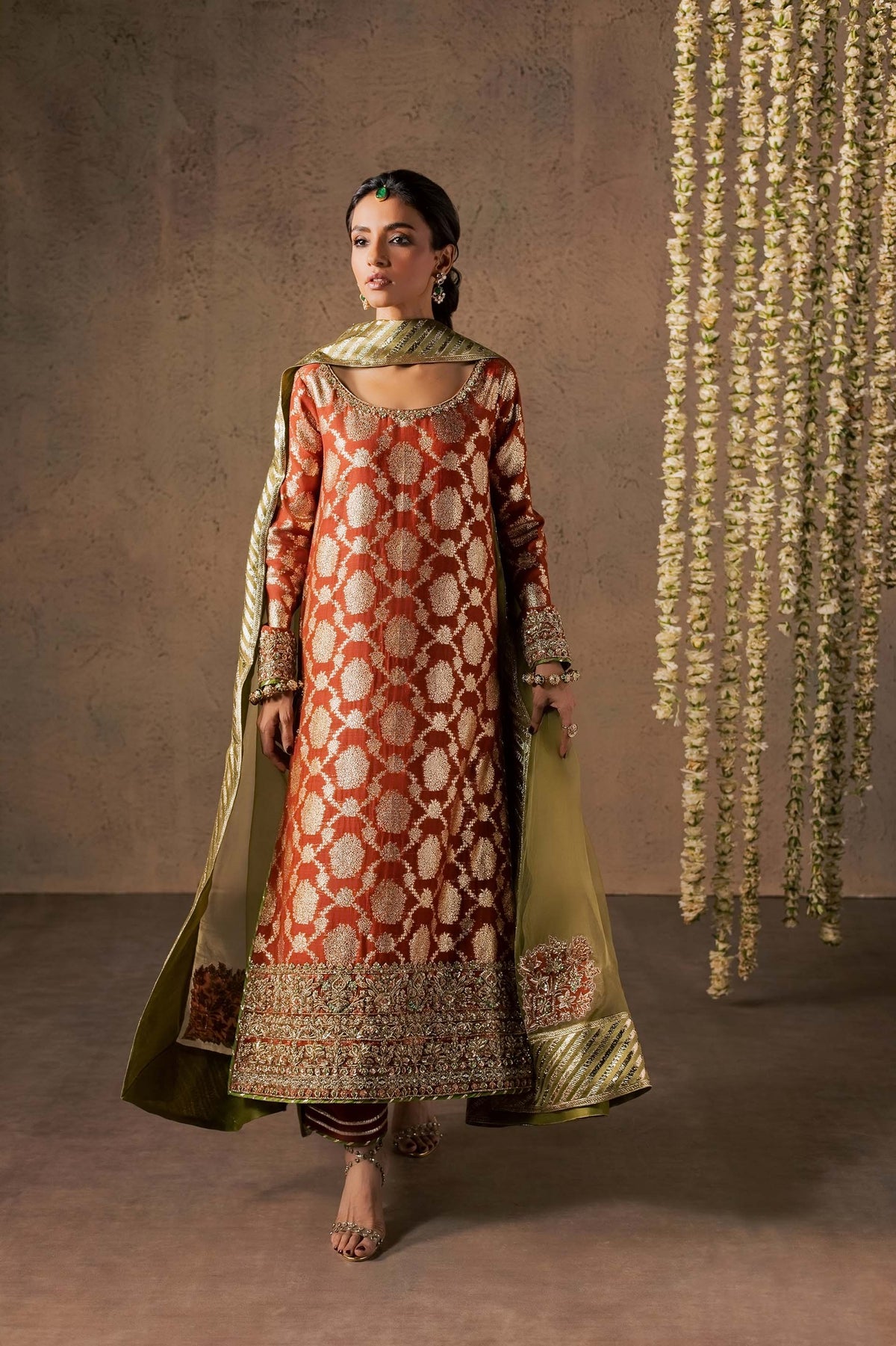 Zonaira Ahmed Mehr rust gold wedding formal 3-piece – full front standing view with the dupatta flowing open, floral garland backdrop