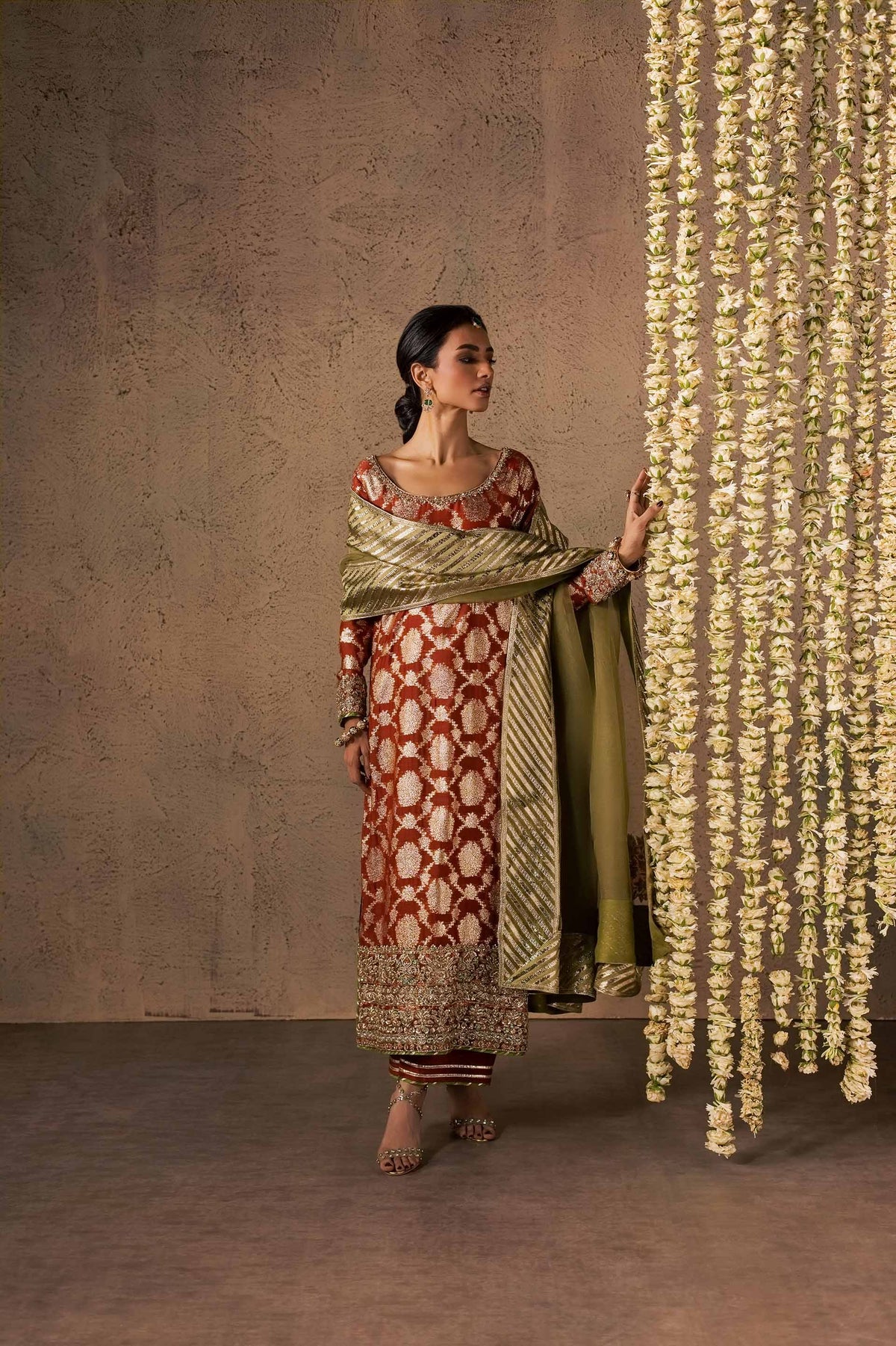 Zonaira Ahmed Mehr rust gold wedding formal 3-piece – full-length lifestyle shot standing beside a hanging floral garland, touching the flowers