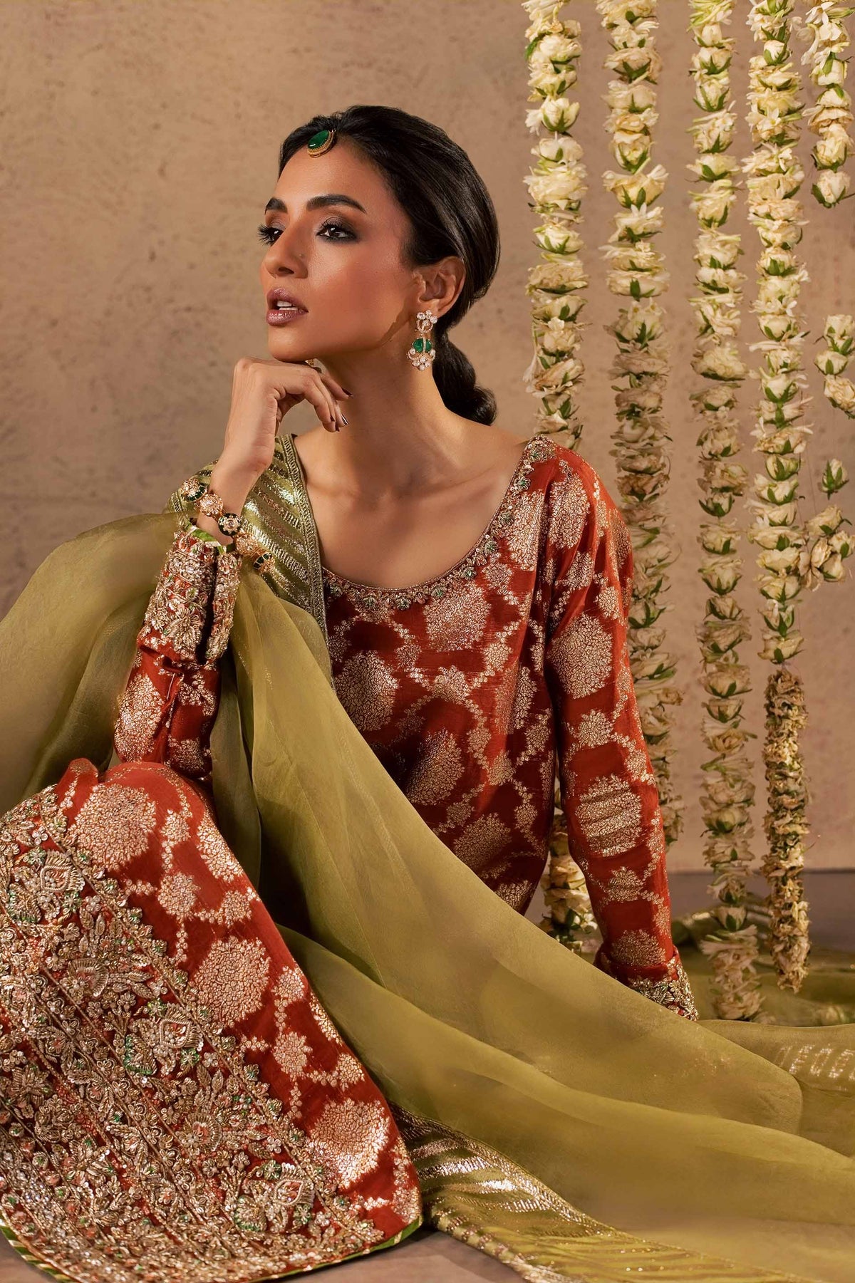 Zonaira Ahmed Mehr rust gold wedding formal 3-piece – seated lifestyle shot with chin resting on hand, floral garland backdrop