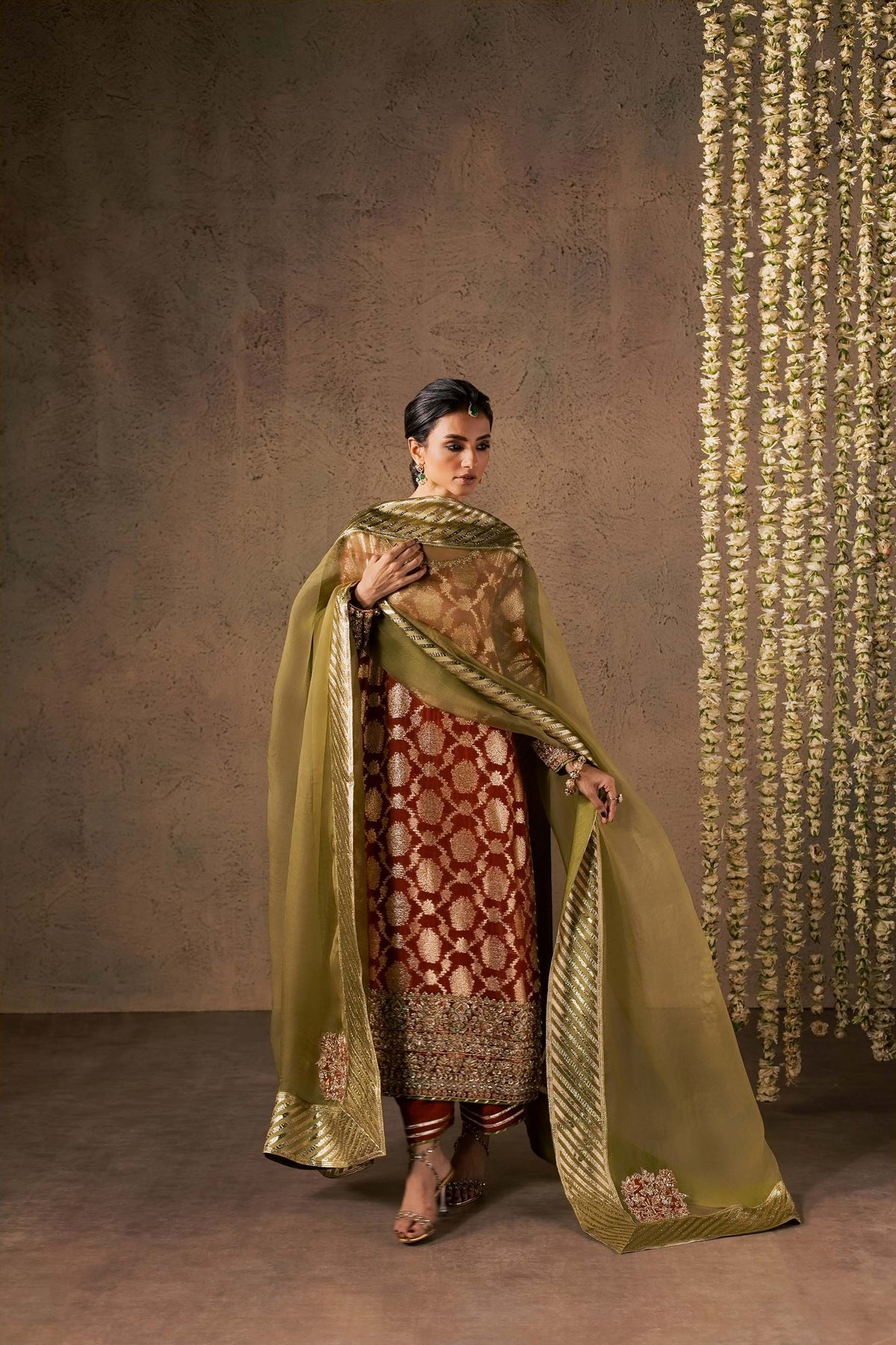 Zonaira Ahmed Mehr rust gold wedding formal 3-piece – full front standing portrait with the striped dupatta draped over both arms, floral garland backdrop