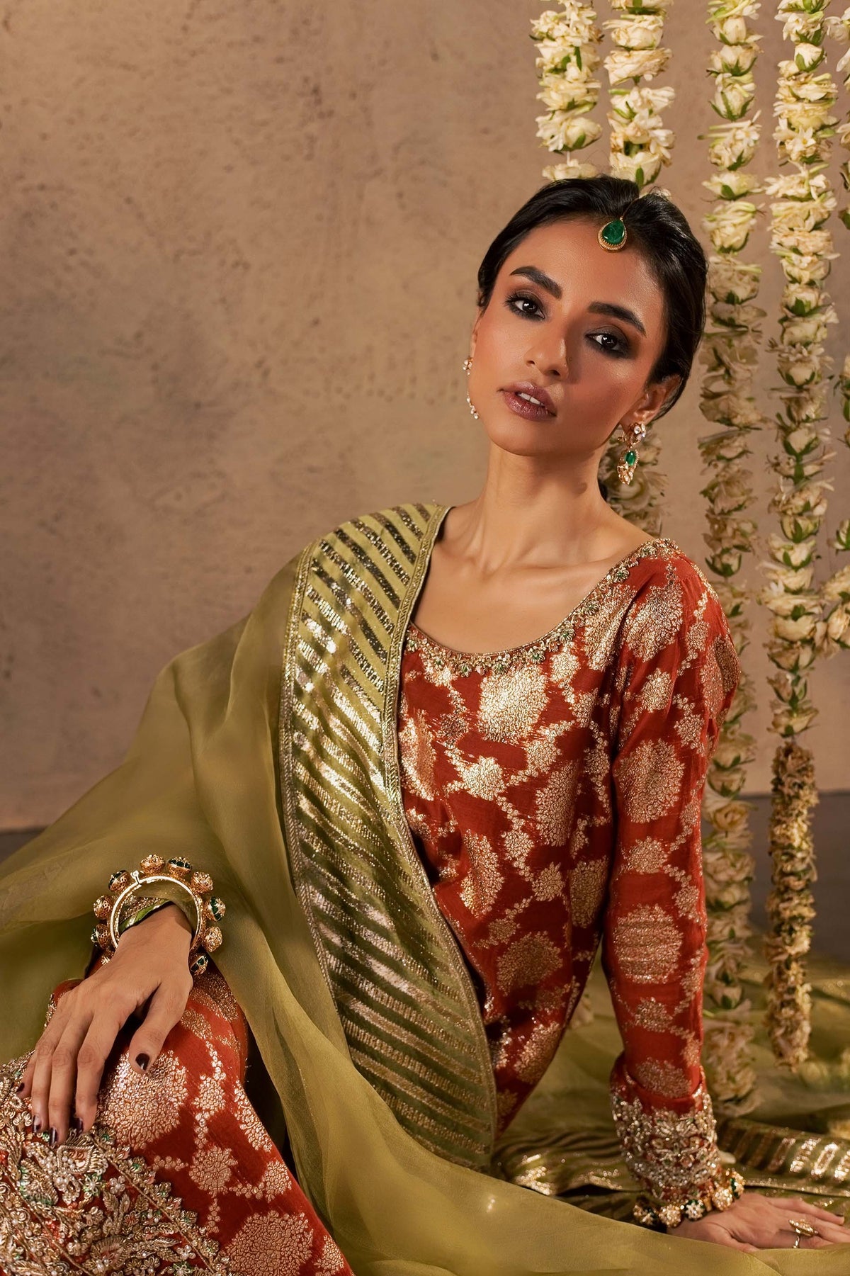 Zonaira Ahmed Mehr rust gold wedding formal 3-piece – seated portrait showing the jeweled bangle and gold-striped dupatta, floral garland backdrop