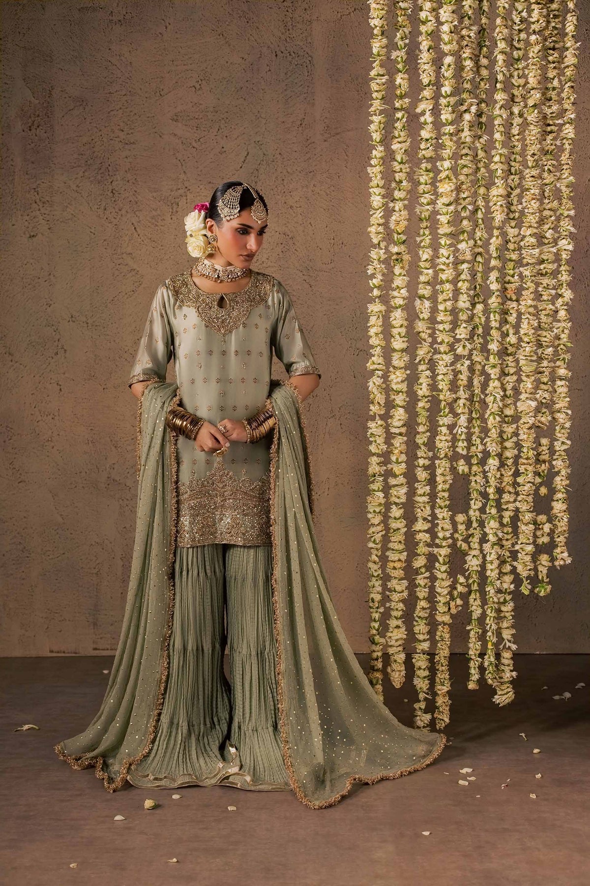 Zonaira Ahmed Naaz sage green wedding formal 3-piece – front-side view with head turned, hands clasped at the waist, floral garland backdrop