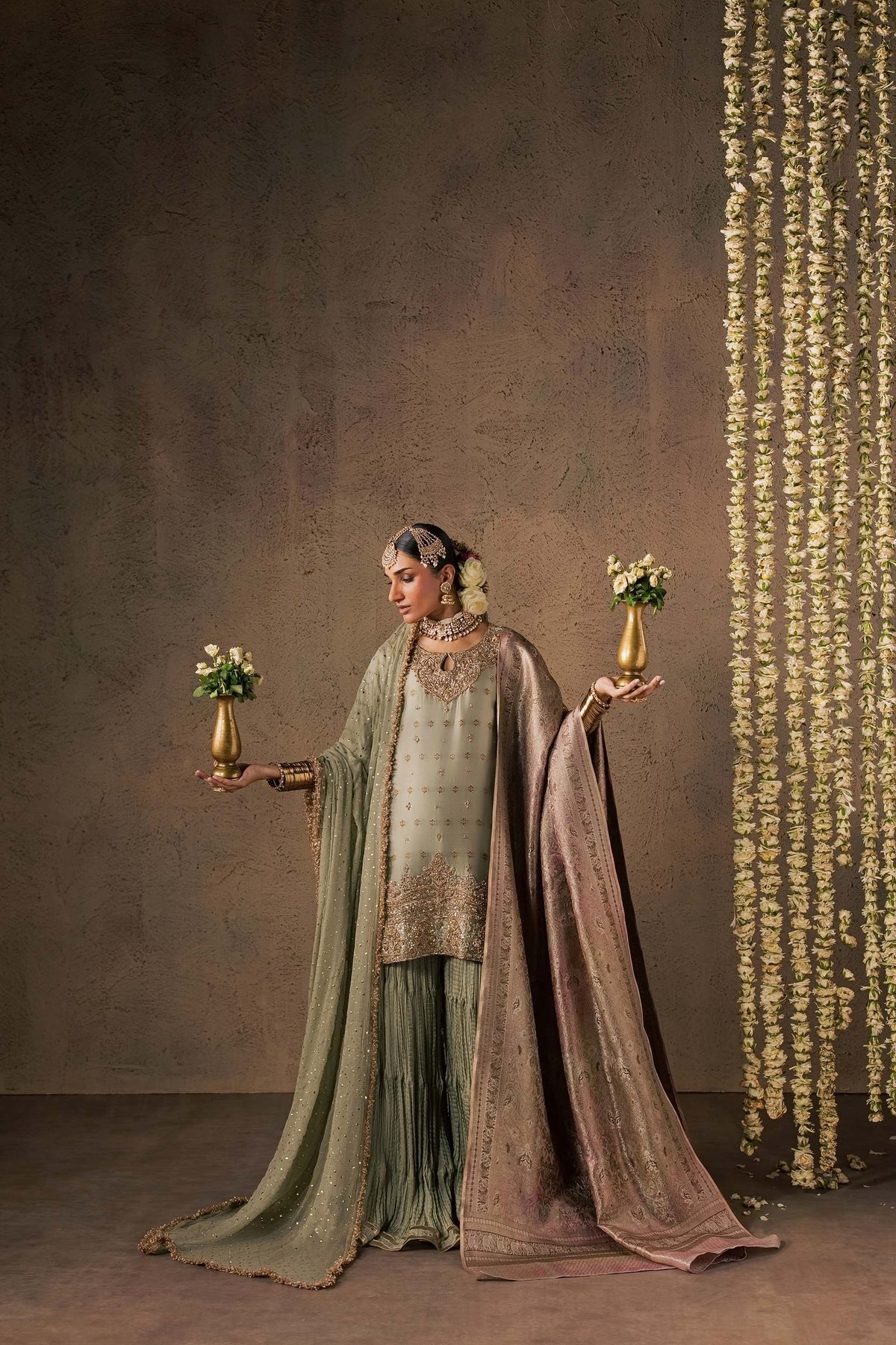 Zonaira Ahmed Naaz sage green wedding formal 3-piece – lifestyle shot holding gold flower vases against floral garlands