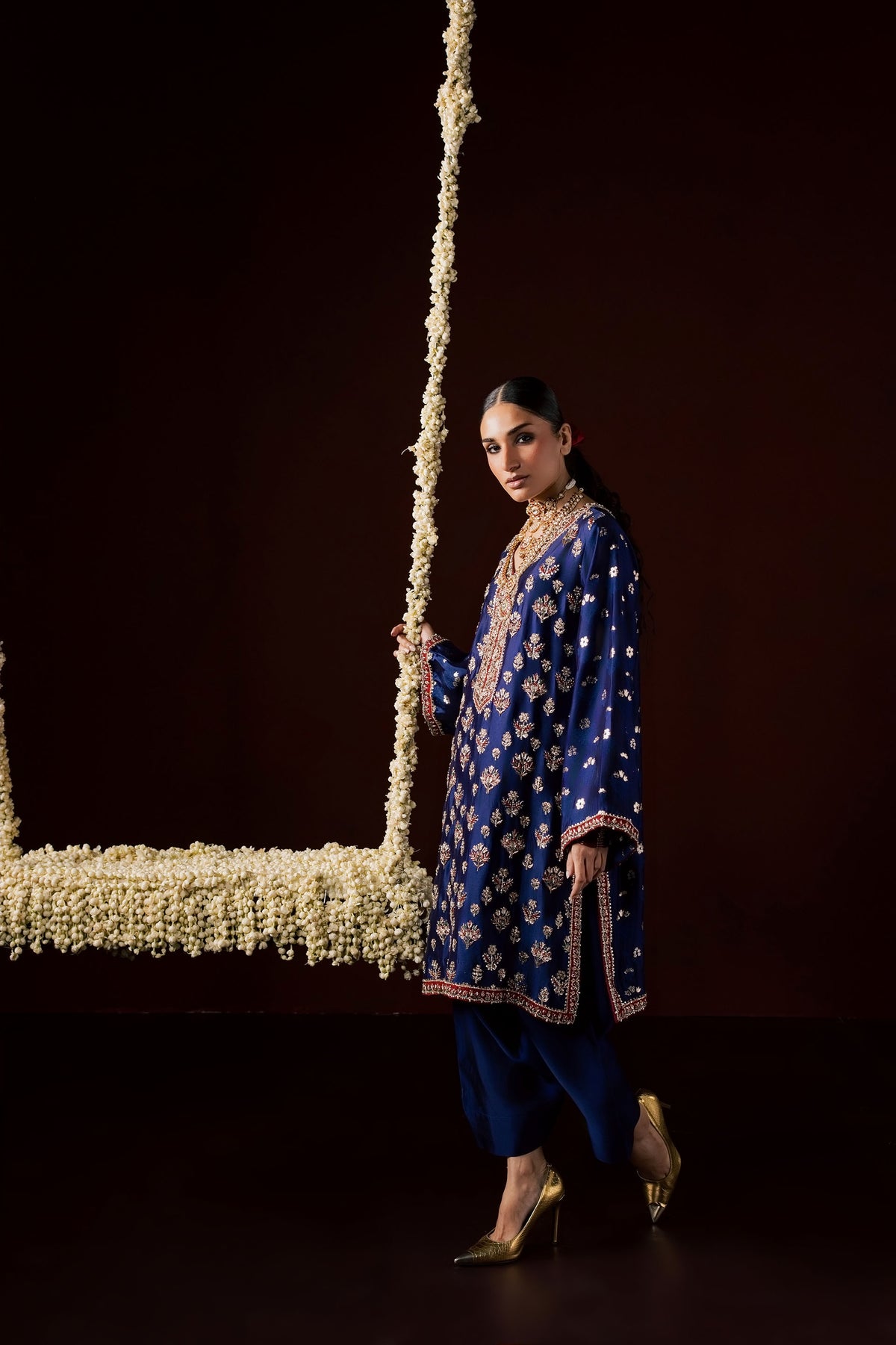 Zonaira Ahmed Nayab navy blue wedding formal 3-piece – three-quarter angle view of the embroidered kurta and shalwar, standing beside a flower-draped swing