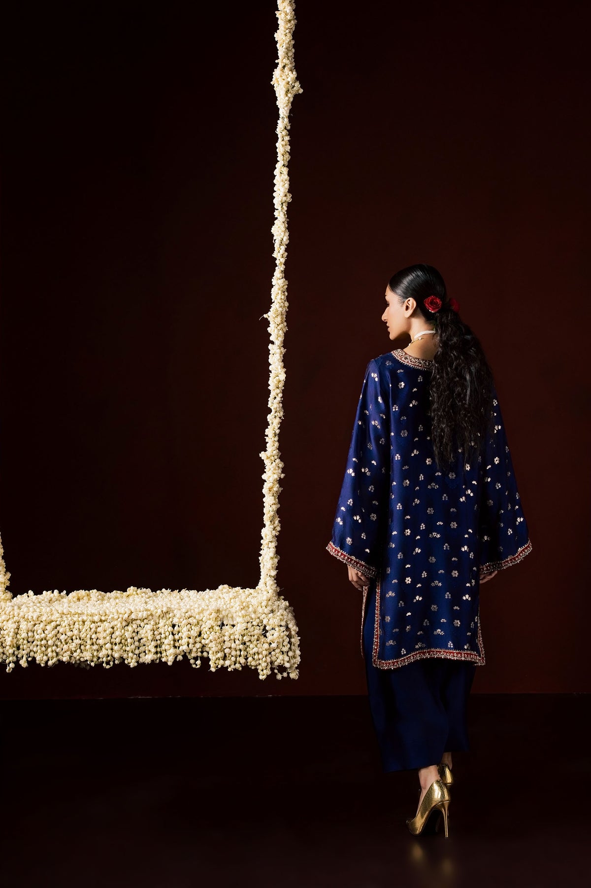 Zonaira Ahmed Nayab navy blue wedding formal 3-piece – back view showing the contrasting red border detail on the kurta, standing beside a floral swing