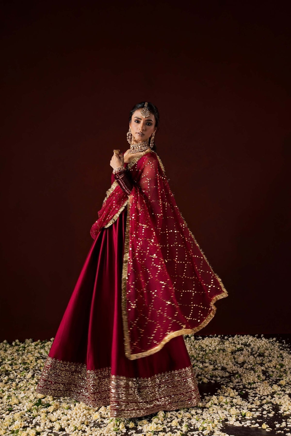 Zonaira Ahmed Neysa maroon wedding formal peshwas – three-quarter angle, dupatta flowing to the side, standing among scattered flower petals