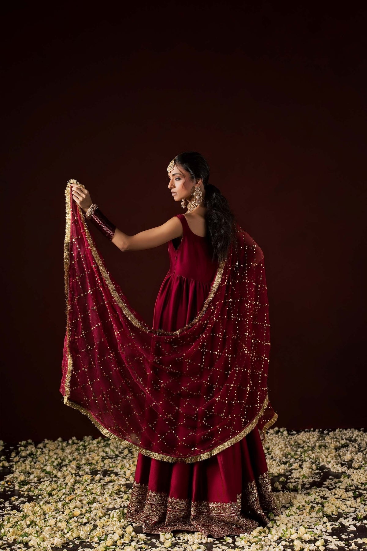 Zonaira Ahmed Neysa maroon wedding formal peshwas – back three-quarter view with the gold mukesh dupatta swirled out, standing among scattered flower petals