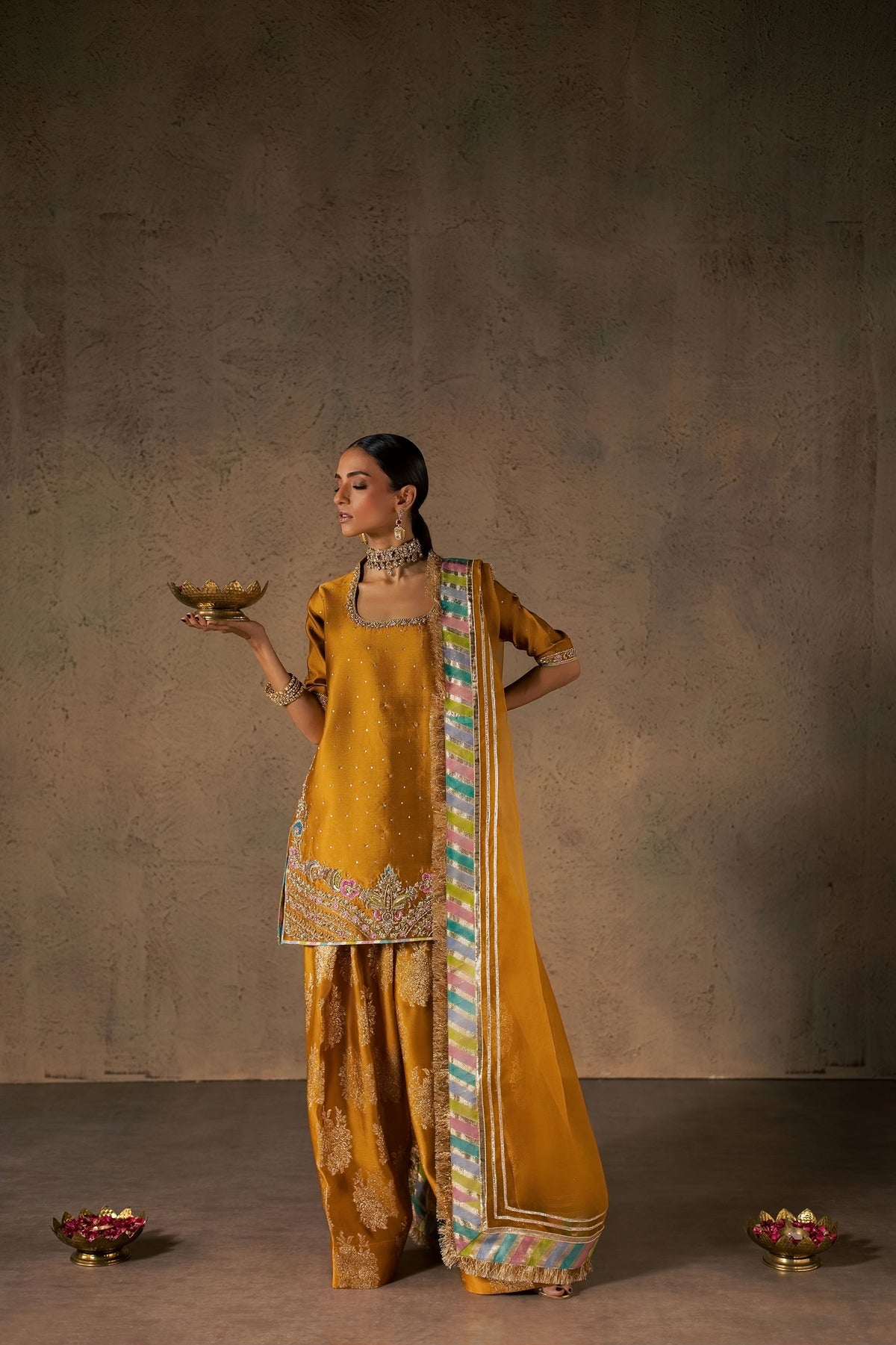 Zonaira Ahmed Zarmin mustard wedding formal 3-piece – front standing view holding a brass diya bowl, hand on hip