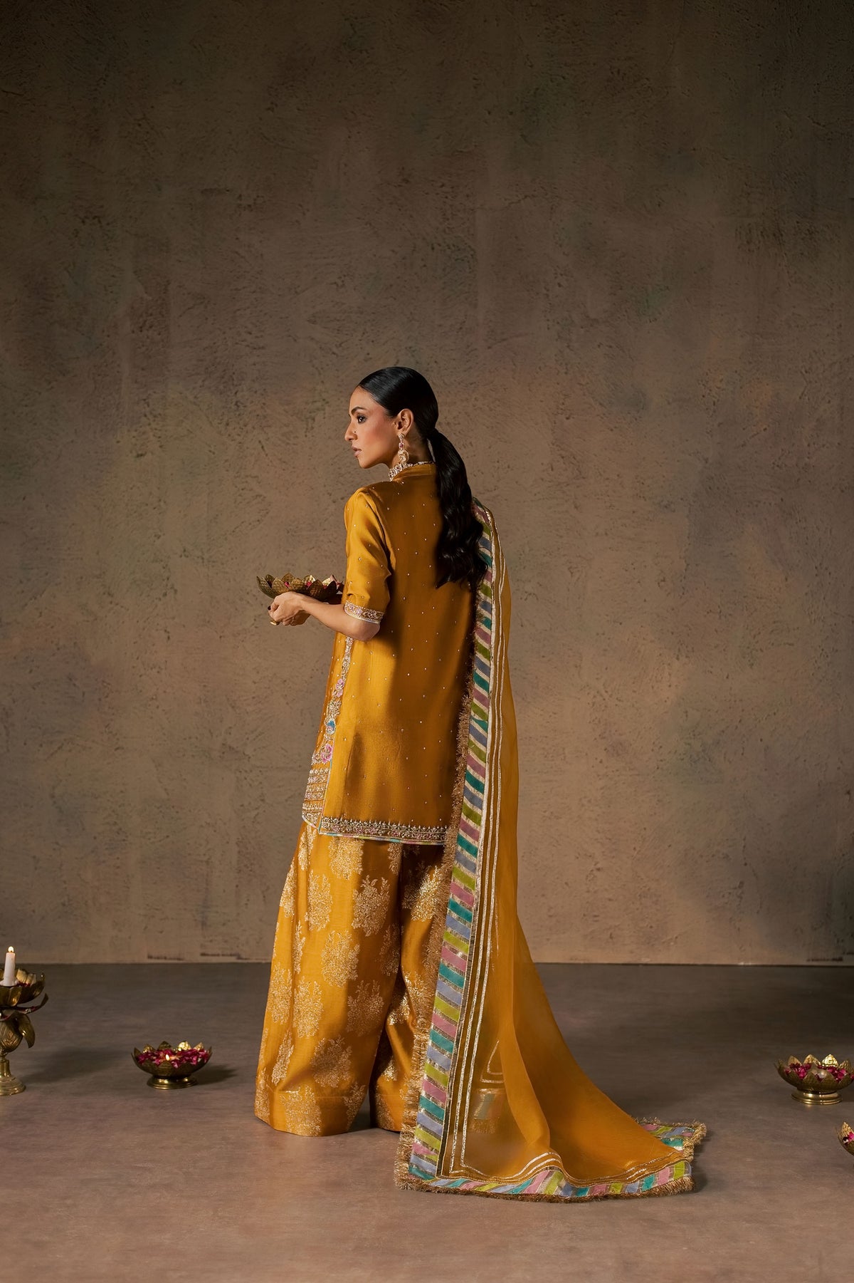 Zonaira Ahmed Zarmin mustard wedding formal 3-piece – back three-quarter view holding a brass diya bowl